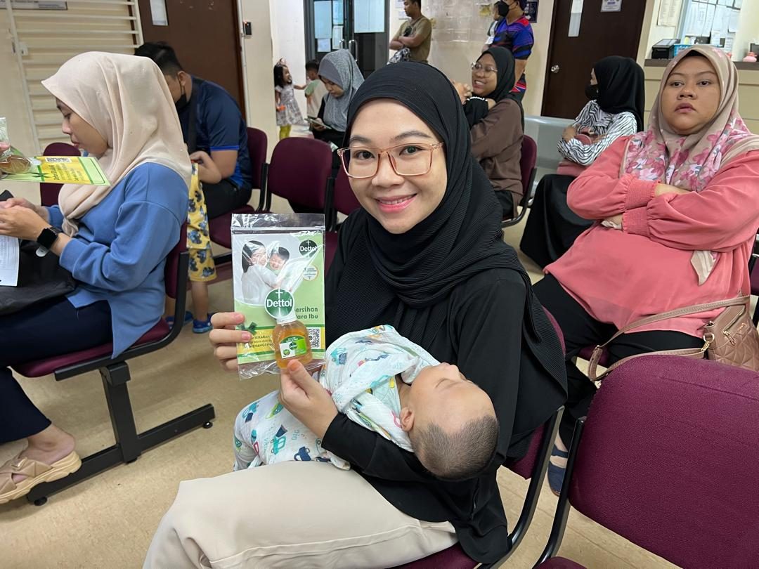 A mother holds her newborn and a Nurture Circle sample pack in a Malaysian clinic waiting area
